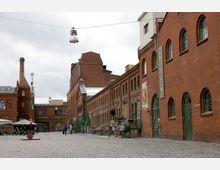 Ein öffentlicher Platz mit Kopfsteinpflaster, umgeben von langen historischen Backsteingebäuden mit bogenförmigen Fenstern; im Hintergrund ein Schornstein und ein Gebäude mit einem Satteldach. Auf dem Platz sind wenige Spaziergänger und vereinzelte Sitzgelegenheiten zu sehen.