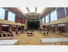 Großer Veranstaltungsraum mit hellem Holzboden, einer Bühne im Hintergrund und einem Dachfenster mit Glaspanelen. Im Raum sind Tische und Stühle in Reihen angeordnet, rote Vorhänge und kugelförmige Hängelampen rahmen die hohen Fenster ein.