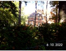 Ein zweistöckiges Gebäude mit roten Wänden und weißen Fensterrahmen steht hinter hohen Bäumen und Büschen in einer bewaldeten Umgebung. Im Vordergrund ist ein Maschendrahtzaun zu sehen.