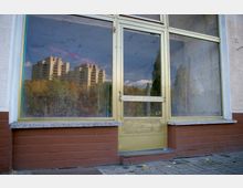 Eingang eines leerstehenden Gebäudes mit großen Glasfenstern und einer gelblichen Metalltür, in der sich Bäume und ein mehrstöckiges Wohngebäude spiegeln. Die Fassade ist teilweise aus roten Ziegelsteinen und Putz gestaltet.