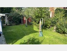 Grüne Gartenfläche mit kurzen Rasen, jungen Bäumen mit Bewässerungssäcken und dichter Bepflanzung im Hintergrund. Rechts ein rotes Gartenhäuschen, links ein überdachter Abstellplatz mit Fahrrädern.