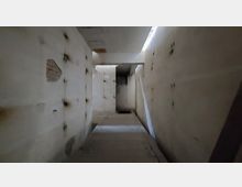 Ein schmaler, abgenutzter Korridor mit schmutzigen weißen Wänden, teilweise mit Löchern und Flecken. Im Hintergrund ist eine kleine Tür sowie eine schwach beleuchtete Betondecke zu sehen.