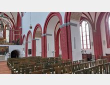 Innenraum einer Kirche mit roten Ziegelpfeilern und gotischen Rundbögen, helle Wände und große Buntglasfenster. Im Vordergrund sind hölzerne Stuhlreihen zu sehen, im Hintergrund befindet sich ein Altarbereich mit Kreuz und Gemälden.