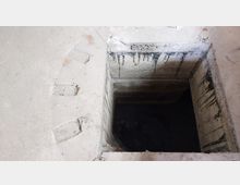 Betonschacht mit rechteckiger Öffnung, umgeben von einer glatten, grauen Betonfläche. Innen sind die Wände des Schachts ebenfalls aus Beton und zeigen Spuren von Feuchtigkeit und Abnutzung.