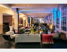 Industriell gestalteter Lounge-Bereich in einem großen Raum mit Sichtbeton-Decke, stilisierten Sofas und Sesseln in verschiedenen Farben, sowie großen Fensterfronten mit Rahmen aus Metall. Die Beleuchtung wirkt stimmungsvoll, mit Stehlampen und dekorativen Hängelampen.