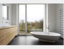 Helles, modernes Badezimmer mit freistehender Badewanne, großem Fenster mit Blick auf eine Landschaft und einem Holzwaschtisch mit Spiegel an der Wand. Ein Heizkörper mit Handtuchhalter ist an der rechten Wand befestigt.