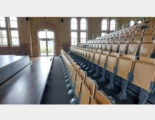 Innenansicht eines Hörsaals mit ansteigenden Sitzreihen aus Holz und Metallgestellen. Der Raum hat große Fenster mit halbrunden Bögen und Wände aus gelbem Backstein, rechts eine Tür mit Blick nach außen.