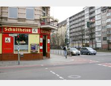 Eckkneipe mit gelb-roter Fassade und dem Schriftzug "Schultheiss" an einer Straßenecke im Stadtteil Alt-Moabit, Berlin. Im Hintergrund sind mehrstöckige Wohngebäude, Autos und eine Fußgängerampel zu sehen.