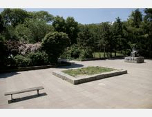 Platz in einem Park mit einer gepflasterten Fläche, einer Bank und einer kleinen, rechteckigen Grünfläche in der Mitte; im Hintergrund dichte Vegetation und rechts eine Statue auf einem Sockel.