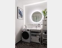 Modernes Badezimmer mit einer weißen Waschmaschine unter einem Waschbecken mit Stauraum darunter. Ein beleuchteter runder Spiegel hängt an der Wand, daneben eine Zimmerpflanze und ein weißer Schrank. Der Boden ist mit dekorativen Fliesen verlegt, und an der Wand hängt ein Landschaftsbild.