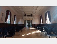 Ein historischer Saal mit hellrosa Wänden und hohen Decken, großen Rundbogenfenstern mit roten Vorhängen, Holztäfelung an den Wänden, einem Holzboden und schwarzen Stühlen, die in Reihen für eine Veranstaltung aufgestellt sind. Im Hintergrund sind Säulen und ein Durchgang zu einem angrenzenden Bereich sichtbar.