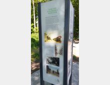 Eine Informationstafel im Freien mit dem Titel „Park und Schlösschen Monplaisir“. Die Tafel zeigt Text und historische Fotos des Parks und Gebäudes, begleitet von einer Beschreibung der Geschichte. Die Tafel steht auf einem gepflasterten Weg, im Hintergrund ist grüne Vegetation sichtbar.