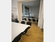 Ein Konferenz- oder Schulungsraum mit Holzboden, mehreren rechteckigen Tischen, schwarzen Stühlen und großen Fenstern mit dunkelgrauen Jalousien. Im Hintergrund steht ein Beamer auf einem Tisch, und hinter einer geöffneten Schiebetür ist ein weiterer Bereich sichtbar.