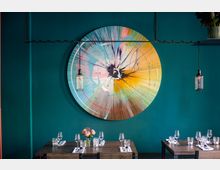 Ein modernes Restaurant mit dunkelgrüner Wand, an der ein großes, buntes, abstraktes Wandbild in Kreisform hängt. Im Vordergrund stehen gedeckte Holztische mit Gläsern, Besteck und Blumen, darüber hängen zwei hängende Glühbirnenlampen.