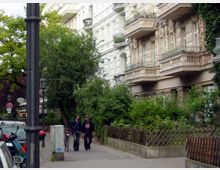Städtische Straßenszene mit historischen Wohngebäuden im Altbaustil, verzierten Balkonen und Rankpflanzen an den Fassaden. Der Bürgersteig ist von Bäumen und kleineren Grünflächen gesäumt, vorbeigehende Menschen sowie parkende Autos und Fahrräder sind sichtbar.