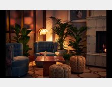Gemütliche Lounge mit blauen Polstersesseln, gerahmt von großen Zimmerpflanzen und einem weißen Kachelofen mit Kaminfeuer. Im Vordergrund stehen ein Holztisch mit Getränken und geflochtene Fußhocker; eine warme Beleuchtung dominiert den Raum.