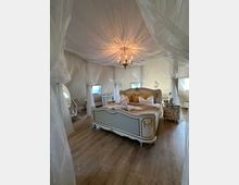 Schlafzimmer mit romantischer Einrichtung, bestehend aus einem verzierten Himmelbett, Holzfußboden, weißen Vorhängen und einer zentralen, dekorativen Deckenleuchte. Im Raum stehen ein antiker Spiegel und passende Beistelltische mit Ornamentdetails.