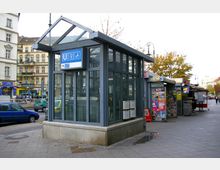 Glasüberdachter Eingang zur U-Bahnlinie U7 mit blauen Schildern und einem Rollstuhlsymbol, umgeben von Kiosken und mehrstöckigen Wohngebäuden in einer städtischen Umgebung. Im Hintergrund sind geparkte Autos und Bäume sichtbar.