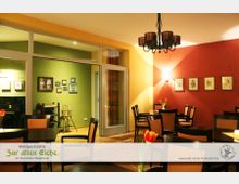 Gemütlicher Gastraum mit roten und gelben Wänden, dekoriert mit gerahmten Bildern und Wandbeleuchtung. Schwarze Holztische und gepolsterte Stühle sind verteilt, und in der Mitte des Raumes hängt ein dunkler Kronleuchter.