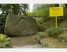 Großer Findlingsstein mit geraden Flächen in einer bewaldeten Umgebung, flankiert von einer gelben Informationstafel mit der Aufschrift "Geologisches Naturdenkmal Kobbelsner Stein". Der Bereich ist von Steinmauern und Pflanzen umgeben.