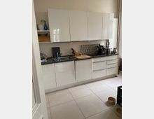 Moderne Küche mit weißen Hochglanzschränken, grauer Arbeitsplatte und hellen Fliesenboden. Neben einer eingelassenen Spüle befinden sich Küchenutensilien sowie ein Holzschneidebrett; Glasbausteine an der Wand dienen als dekoratives Element.