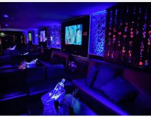 Ein moderner Clubraum mit dunklen Ledersofas, beleuchtet durch blau-lilane Akzente. An den Wänden befinden sich abstrakte Spiegelmuster, Fernseher mit Bildschirmen und glitzernde Dekoelemente aus Perlen.