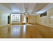 Großer, leerer Raum mit hellen Wänden aus glänzenden Beigen Ziegeln, einem Holzparkettboden und einer dekorativen Deckenleuchte. Große Fenster lassen Tageslicht herein, eine kleine Treppe führt zu einer offenen Tür, die nach außen zeigt.