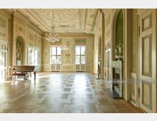 Ein prunkvoller, historischer Saal mit reich verzierten Wänden und Decken im klassizistischen Stil, zwei großen Fenstern, einem Kronleuchter und einer hellen Parkettbodenfläche; links steht ein Klavier, rechts befinden sich ein Kamin und verzierte Türen.