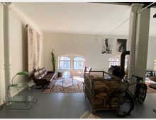 Ein heller Raum mit minimalistischer Einrichtung, darunter eine braune Ledercouch, ein Glastisch mit grünem Lampenschirm und ein Holzofen mit einem Korb voller Holz. Große Rundbogenfenster lassen Tageslicht herein, und an den Wänden hängen schwarz-weiße Fotografieposter.