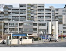 Wohnkomplex mit moderner, funktionaler Architektur, bestehend aus mehreren Etagen mit Balkonen und großen Fenstern. Im Vordergrund befindet sich der U-Bahn-Eingang "U-Bhf Richard-Wagner-Platz" mit umliegenden Geschäften und Verkehrsschildern.