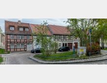 Kopfsteinpflasterstraße mit einer Reihe von Fachwerkhäusern, die rote Ziegeldächer und helle Fassaden haben. Im Vordergrund sind Bäume, Sträucher und eine kleine Grünfläche mit parkenden Autos zu sehen.