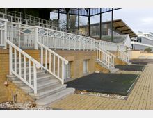 Außenansicht einer Tribüne aus gelbem Klinker mit mehreren Treppenaufgängen, weißen Geländern und überdachten Sitzbereichen. Im Hintergrund sind moderne Glas- und Metallstrukturen zu sehen.