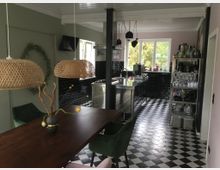 Offene Wohnküche mit modernem Design. Ein Essbereich mit dunklem Holztisch, grünen Polsterstühlen und geflochtenen Hängelampen grenzt an eine Küche mit schwarz-weißen Schachbrettfliesen, schwarzen Wandfliesen, einer Edelstahlinsel und großen Fenstern mit Blick ins Grüne.