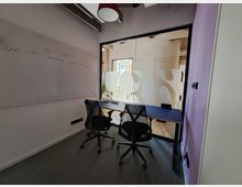 Ein kleiner, moderner Büroraum mit zwei schwarzen Drehstühlen, einem Tisch und einer großen Glaswand mit abstrakten weißen Mustern. An der linken Wand befindet sich ein beschreibbarer Whiteboard-Bereich mit handschriftlichen Notizen.