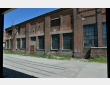 Backsteinfassade eines alten Industriegebäudes mit großen, rechteckigen Fensterreihen und einer kleinen Holztür. Vor dem Gebäude sind Schienen und ein gepflasterter Weg zu sehen, umgeben von vereinzeltem Grasbewuchs.