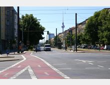 Straßenszene in einer Stadt mit breiter Kreuzung, roten Radwegen und Straßenbahnschienen in der Mitte. Im Hintergrund sind Wohnhäuser, Bäume und der Berliner Fernsehturm zu sehen.