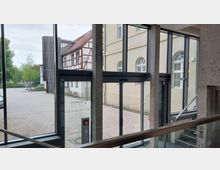 Blick aus einem modernen Gebäudeeingang mit breiten Glastüren und Betonpfeilern auf einen Innenhof. Im Hintergrund sind ein Fachwerkgebäude sowie ein gelbes Gebäude mit Rundbogenfenstern zu sehen, umgeben von Pflasterflächen und Grünbereichen.
