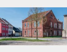 Backsteinbau im Stil der alten Industriearchitektur mit zugemauerten Fenstern und Türen, umgeben von weiteren Häusern aus unterschiedlichen Bauzeiten, darunter ein rotes Wohnhaus. Im Vordergrund ist eine Wiese, ein Baum und eine gepflasterte Gehwegfläche sichtbar.