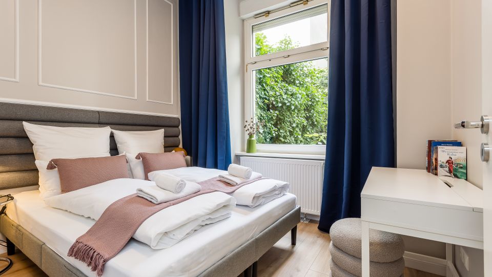 Helles Schlafzimmer mit Doppelbett, gepolstertem Kopfteil und weißer Bettwäsche, dazu ein kleiner weißer Schreibtisch und Hocker. Großes Fenster mit dunkelblauen Vorhängen und Blick ins Grüne, darunter ein Heizkörper.