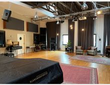 Ein geräumiger Musik- oder Aufnahmestudio-Raum mit Holzboden, großen grauen Vorhängen und mehreren Teppichen. Der Raum enthält Instrumente wie ein Klavier, Audiotechnik, gemütliche Sessel und Deckenleuchten an freiliegenden Holzbalken.