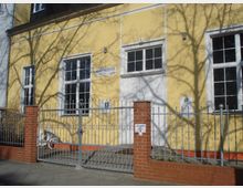 Gelbes Gebäude mit hohen weißen Fenstern und einer weißen Tür, umgeben von einem silbernen Zaun mit roten Backsteinpfosten. Im Vordergrund steht ein Schild mit Kontaktdaten und ein Baum wirft Schatten auf die Fassade.