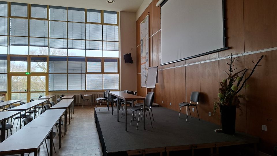 Großer Seminar- oder Konferenzraum mit hohen, rasterförmigen Fensterflächen und Jalousien sowie Reihen aus Tischen und Stühlen. Rechts befindet sich eine erhöhte Bühne mit Projektionsleinwand, Holzvertäfelung an der Wand und einzelnen Stühlen sowie einer Pflanzendekoration.