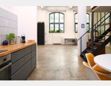 Heller, loftartiger Innenraum mit offener Küchenzeile links, Betonboden und weißen Wänden mit großen, bogenförmigen Sprossenfenstern. Rechts führt eine schwarze Metalltreppe an einer Glaswand vorbei; im Vordergrund steht ein runder Tisch mit Stühlen.