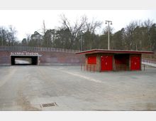 Backsteinunterführung mit der Aufschrift "Zum Olympia-Stadion" in einem offenen, gepflasterten Bereich; rechts im Vordergrund ein kleines Gebäude mit Flachdach und roten Türen. Im Hintergrund kahle Bäume und ein trüber Himmel.