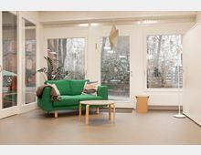 Heller Aufenthaltsraum mit großem Fenster- und Türelement zur Außenseite, grauem Boden und weißer Decke. In der Mitte stehen ein grünes Sofa mit Kissen, ein runder Couchtisch sowie eine Stehlampe und eine Hängelampe; draußen sind Bäume und Nachbargebäude zu sehen.