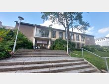 Außenansicht eines zweigeschossigen Gebäudes mit großen Fenstern und flachem Dach, umgeben von Bäumen und Sträuchern. Eine Treppe mit Geländern führt zum Eingang, über dem ein Schild mit der Aufschrift „Campus für Demokratie“ angebracht ist.