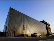 Ein modernes Industriegebäude mit glatter, metallischer Außenfassade, im Sonnenlicht glänzend. Im Vordergrund steht ein geparktes Auto, und daneben sind kleinere Bäume entlang eines Gehwegs zu sehen.
