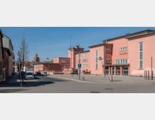 Straßenszene mit dem rosa Gebäude eines Stadttheaters im Bauhaus-Stil, flankiert von weiteren Gebäuden und einer Kirche mit Turm im Hintergrund. Im Vordergrund sind parkende Autos, Straßenlaternen und Verkehrszeichen sichtbar.
