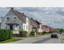 Straßenansicht eines Wohnviertels mit freistehenden Einfamilienhäusern, die teilweise verputzt oder gestrichen sind, und Satteldächern. Im Vordergrund fährt ein Motorrad mit Beiwagen die Straße entlang, während Gehwege, Laternen und Gartenanlagen die Bebauung ergänzen.