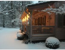 Holzhaus mit beleuchteter Veranda in einer verschneiten Waldlandschaft. Die Umgebung ist von Schnee bedeckt, und kahle Äste tragen eine Schicht Schnee.